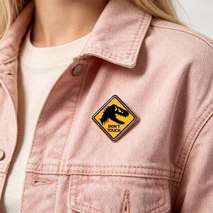 May include: A diamond-shaped enamel pin featuring a yellow background with a black silhouette of a dinosaur head and the text DON'T TOUCH in black lettering, attached to a light pink denim jacket worn over a cream-colored shirt.