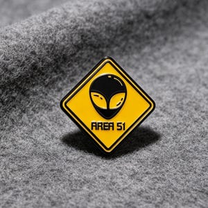 Peut inclure: Une broche en émail en forme de losange avec un fond jaune et une bordure noire. Le motif présente un graphique de tête d'extraterrestre noir centré au-dessus du texte noir AREA 51, imitant l'apparence d'un panneau de signalisation routière.
