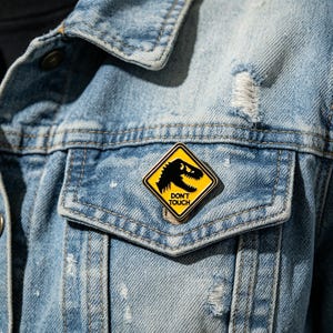 May include: A diamond-shaped enamel pin attached to a light blue distressed denim jacket. The pin features a yellow background with a black silhouette of a dinosaur head and the text DON'T TOUCH printed in black capital letters on the lower half.