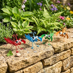 May include: Four metal insect sculptures in red, blue, green, and orange. Each figure features large white eyes with black pupils, thin wire legs, and curved pincers. The insects are arranged in a row, standing on a textured stone surface.