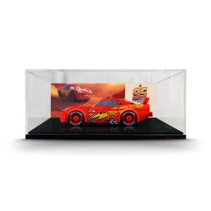 May include: A red toy race car model featuring the number 95 and a yellow lightning bolt graphic on the side, displayed inside a clear rectangular acrylic case with a black base and a printed background showing a desert racing scene.