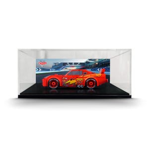 May include: A red toy race car with the number 95 and a yellow lightning bolt graphic on the side, enclosed in a clear rectangular display case. The car features black wheels with white lettering and is set against a racetrack background.