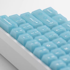 May include: A mechanical keyboard featuring translucent, light blue keycaps with white lettering. The keys display standard symbols and text including Backspace, Enter, Shift, Fn, and Super. The keyboard has a white base and a compact layout.