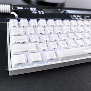 Könnte beinhalten: Eine mechanische Tastatur mit weißen Tastenkappen, schwarzer Beschriftung und einem transparenten Außenrand. Die Tastatur hat einen weißen Rahmen und ist über ein weißes Spiralkabel verbunden. Die Tasten zeigen ein Standard-QWERTY-Layout mit Beschriftungen wie Tab, Caps Lock, Shift, Ctrl, Super und Alt.