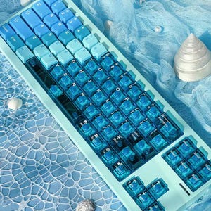 Könnte beinhalten: Eine mechanische Tastatur mit einem hellblauen Rahmen und einem Farbverlauf aus blauen Tastenkappen. Die linke Seite hat solide blaue Tasten, während die rechte Seite und die Navigationstasten aus transparentem blauem Kunststoff bestehen.