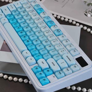 May include: A mechanical keyboard with a white frame and a gradient of blue keycaps. The keys feature standard QWERTY lettering, some with small printed facial expressions and symbols. A small black rectangular display is located on the right side.