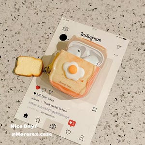 May include: A silicone protective case for wireless earbuds designed to look like a slice of toast topped with a fried egg. The case features the text Yummy Toast and includes a small toast-shaped charm attached to the side by a metal ring.