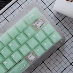 Könnte beinhalten: Eine mechanische Tastatur mit einem durchscheinenden, mattierten Rahmen und hellen mintgrünen Tastenkappen. Die Tasten haben weiße Beschriftungen, darunter Buchstaben, Satzzeichen und Richtungspfeile. Zwei mechanische Schalter sind sichtbar.