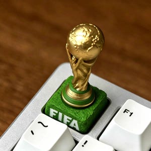 May include: A custom mechanical keyboard keycap featuring a miniature gold-colored FIFA World Cup trophy mounted on a textured green base that resembles a soccer field, with the white text FIFA printed clearly on the front side of the keycap.