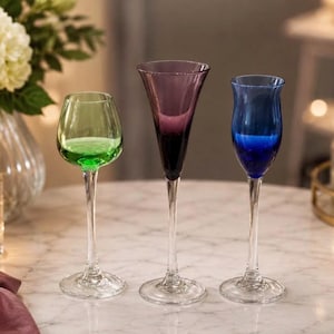 May include: Three stemmed glasses with clear glass bases and stems. The left glass has a rounded green bowl, the center glass features a tall, slender purple flute shape, and the right glass has a tulip-shaped blue bowl. All glasses are transparent.