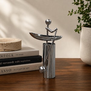 May include: A polished silver-toned metal sculpture depicting a stylized figure sitting in a small boat, holding a long oar that extends downward. The figure is composed of simple geometric shapes, including a spherical head and a cylindrical base.