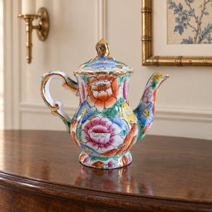 May include: A porcelain teapot featuring a vibrant floral pattern with orange, pink, yellow, and blue flowers on a white background. The teapot includes a matching lid with a gold-trimmed finial, a curved handle, and a spout, all accented with gold.