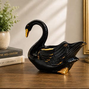 May include: A glossy black ceramic swan figurine featuring gold-painted accents on its beak, wing feathers, and webbed foot. The swan is sculpted in a swimming position with its neck curved gracefully, serving as a decorative bowl or planter.