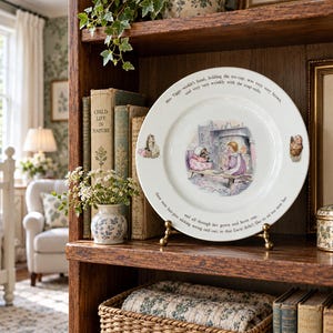 May include: A white ceramic decorative plate featuring a central illustration of Mrs. Tiggy-winkle and Lucie having tea. The rim displays printed text from the Beatrix Potter story, with small character accents on the left and right sides.