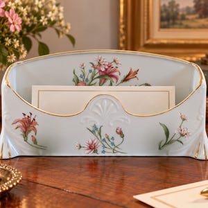 May include: A pale blue porcelain letter holder featuring a delicate floral pattern with pink, red, and green botanical illustrations. The piece has a scalloped front edge and is finished with a thin gold trim along the top and base edges.