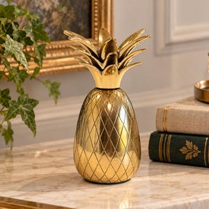 May include: A polished gold-colored metal pineapple figurine featuring a textured diamond-patterned body and a multi-layered pointed leaf top. The object has a reflective, smooth surface finish and stands upright as a decorative tabletop ornament.