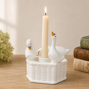 May include: A white ceramic candle holder shaped like a woven basket with three goose figurines attached to the rim. The center features a cylindrical socket holding a single, tall, cream-colored taper candle with a lit flame at the top.