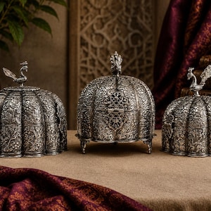 May include: Three silver-toned metal containers with intricate floral engravings and pumpkin-shaped bodies. Each container features a lid topped with a small bird figurine. The central container stands on three small legs, while the two side containers have a flat base.