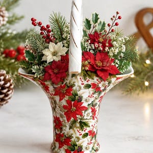May include: A ceramic basket with a flared rim and a white handle, decorated with a pattern of red and white poinsettias and green leaves. The basket is filled with an arrangement of artificial red poinsettias, holly, pine, and red berries.