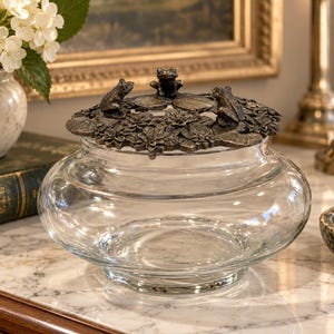 May include: A clear glass bowl featuring a decorative metal lid with a dark bronze finish. The lid is adorned with sculpted lily pads, water lily flowers, and three small frog figures positioned around the circular top edge.