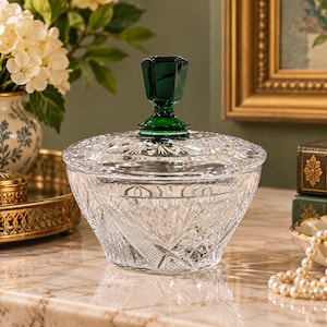 May include: A clear cut glass bowl with a matching lid featuring a deep emerald green faceted finial. The bowl and lid are decorated with intricate geometric starburst and crosshatch patterns that catch the light, creating a brilliant display.