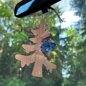 Puede incluir: Un ambientador de coche de madera con forma de pino, con un acabado de veta de madera marrón claro. Una pequeña botella de vidrio facetada en forma de diamante, llena de un líquido azul vibrante, está unida al árbol mediante un cordel fino de color natural.