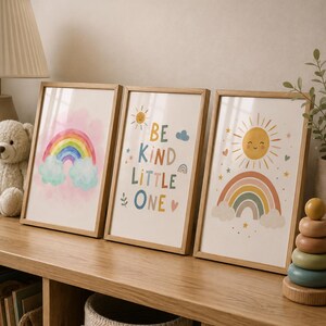 May include: Three rectangular wall art prints in light wood frames. The left print shows a colourful rainbow over clouds. The centre print features the text BE KIND LITTLE ONE with sun, cloud, and heart icons. The right print displays a smiling sun.