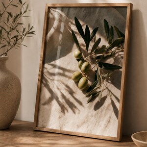 Könnte beinhalten: Ein rechteckiger Wandkunstdruck in einem hellbraunen Holzrahmen mit einem fotografischen Bild eines grünen Olivenzweigs mit mehreren Oliven vor einem strukturierten beigen Hintergrund, dargestellt mit natürlichem Licht und Schatten.
