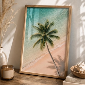 May include: A rectangular framed wall art print featuring an aerial view of a single palm tree on a sandy beach next to turquoise ocean water. The image shows the palm tree casting a long shadow across the light tan sand and clear water.