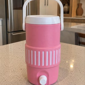 May include: A pink beverage cooler with a white lid and a white handle. The cylindrical container features a textured middle section with vertical white stripes and a white spigot at the bottom for dispensing liquids. The design is simple and functional.