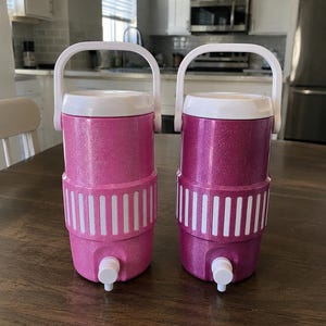 May include: Two cylindrical beverage coolers with white handles and spigots. The left cooler is light pink with a glitter finish, and the right cooler is a darker magenta with a glitter finish. Both feature a white ribbed band around the center.