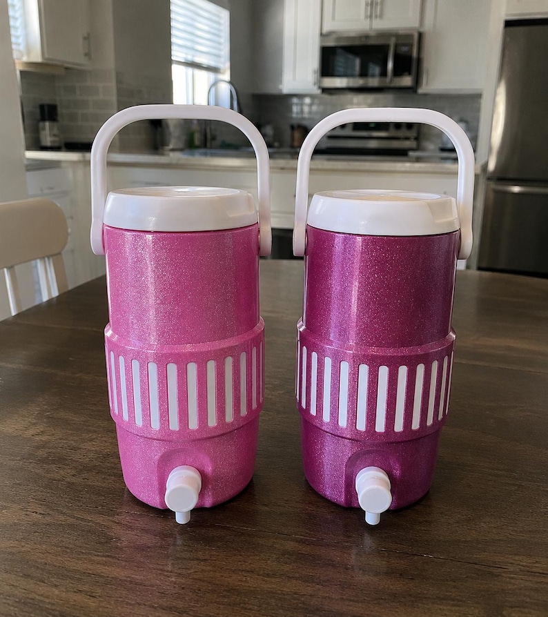 May include: Two cylindrical beverage coolers with white handles and spigots. The left cooler is light pink with a glitter finish, and the right cooler is a darker magenta with a glitter finish. Both feature a white ribbed band around the center.
