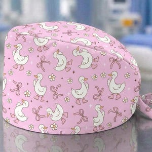 Può includere: Una cuffia chirurgica rosa con un motivo ripetuto di anatre bianche che indossano stivali da pioggia rosa, intervallate da piccoli fiori gialli, nastri rosa e minuscole stelle a quattro punte, con chiusura a laccetti sul retro.