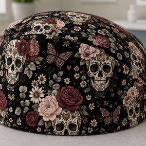 Op de afbeelding: Een zwarte operatiemuts met een herhalend patroon van witte schedels met decoratieve hartvormige details op het voorhoofd, afgewisseld met donkerrode en lichtroze rozen, kleine witte bloemen, groene bladeren en bruine vlinders, met een striksluiting.