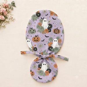 May include: A lavender scrub cap featuring a repeating pattern of white ghosts, orange pumpkins, and green and orange flowers. The fabric is adorned with small black bats and four-pointed stars. The cap includes two fabric ties for securing the fit.