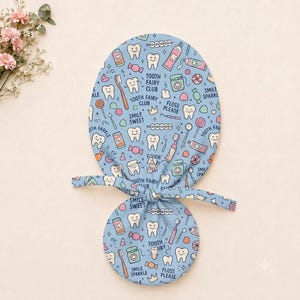 Könnte beinhalten: Eine hellblaue OP-Haube mit einem wiederkehrenden Muster aus weißen Zähnen, rosa Zahnpastatuben, Zahnbürsten, Zahnseide, Bonbons und Kronen. Der Text lautet Tooth Fairy Club, Smile Sweet, Floss Please und Smile Sparkle in dunkelblau.