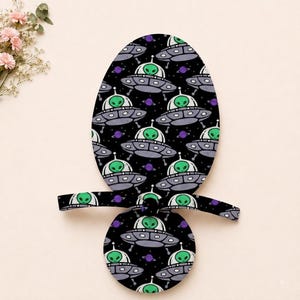 May include: A pacifier featuring a black background with a repeating pattern of grey flying saucers, green aliens, and small purple planets. The design covers both the oval shield and the circular button, with a black strap extending horizontally.