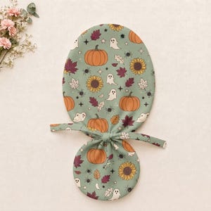May include: A sage green fabric hair accessory featuring a repeating pattern of orange pumpkins, yellow sunflowers, white ghosts, dark red autumn leaves, black spiders, and small stars. The accessory has a rounded top and a smaller circular base.