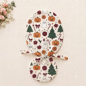 May include: A fabric scrub cap featuring a repeating pattern of orange jack-o'-lanterns, white ghosts, green Christmas trees, red bows, candy canes, and dark red flowers on a cream background, with two fabric ties at the center for adjustment.