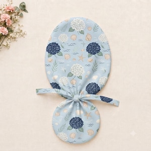 May include: A light blue fabric hair accessory with a pattern of white and dark blue hydrangea flowers, seashells, starfish, and green leaves. The accessory features a rounded top section and a smaller circular base connected by fabric ties.