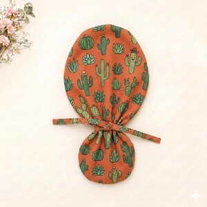 Puede incluir: Un gorro quirúrgico de color óxido con un patrón repetitivo de cactus verdes, algunos de los cuales llevan pequeños sombreros marrones. El gorro tiene una parte superior redondeada y una sección inferior fruncida con dos cintas anudadas.