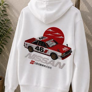 Peut inclure: Un sweat-shirt à capuche blanc avec un motif au dos représentant une voiture de course Nissan rouge et blanche portant le numéro 46. Le design inclut un grand cercle rouge, le texte NISSAN et les caractères japonais 日産自動車株式会社.