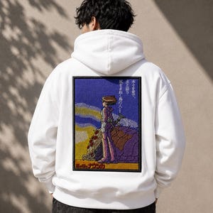 May include: A white hooded sweatshirt featuring a large rectangular embroidered patch on the back. The patch depicts a character standing in a landscape with Japanese text, rendered in shades of purple, yellow, and blue with a black border.