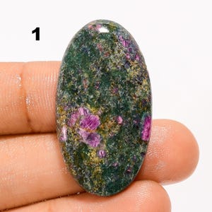 Puede incluir: Una piedra preciosa cabujón de forma ovalada con una base moteada de color verde oscuro y verde bosque, con inclusiones cristalinas de color magenta y rosa vibrante incrustadas por toda la superficie. El número 1 está impreso en negro arriba a la izquierda.