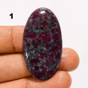 Peut inclure: Un cabochon de pierre précieuse de forme ovale présentant une surface polie avec un motif tacheté d'inclusions magenta profond et gris foncé. La pierre présente un dessus lisse et arrondi ainsi qu'une finition brillante, avec le chiffre 1 visible.