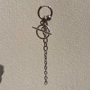 May include: A silver-toned hoop earring featuring a dangling charm assembly. The charm consists of a small metal sphere, a circular ring, a horizontal bar, and a vertical chain link segment, all connected to the main hoop by small jump rings.