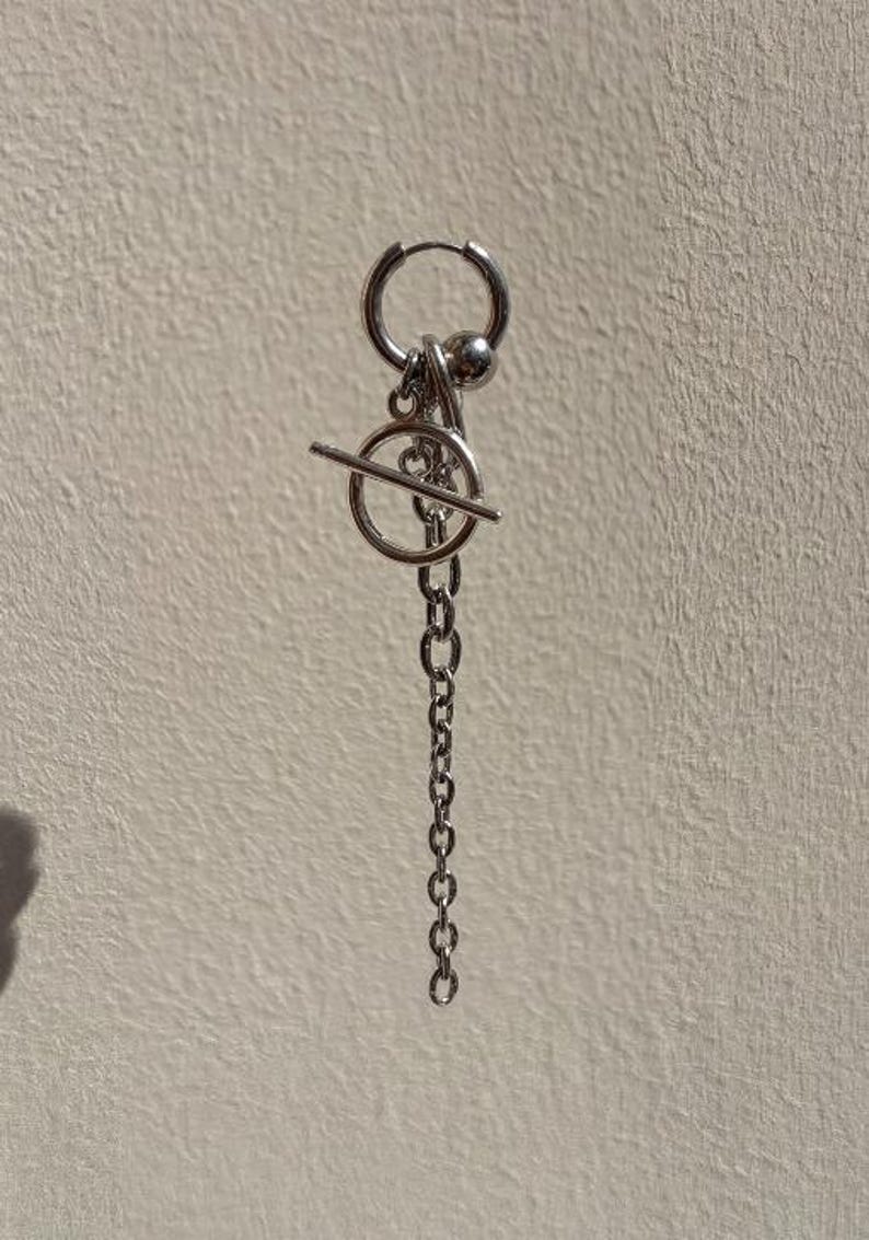 May include: A silver-toned hoop earring featuring a dangling charm assembly. The charm consists of a small metal sphere, a circular ring, a horizontal bar, and a vertical chain link segment, all connected to the main hoop by small jump rings.