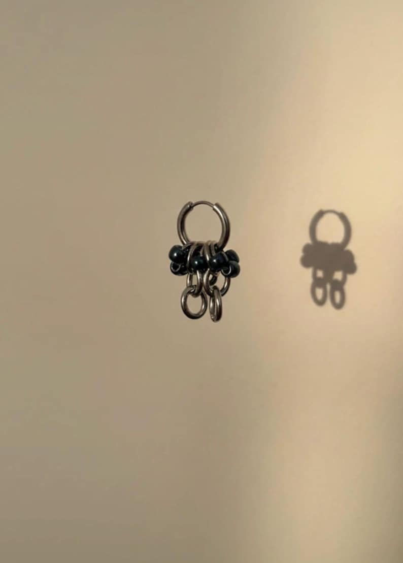 May include: A small hoop earring featuring a series of dangling silver-toned metal rings and dark, round beads. The earring is constructed with a hinged closure mechanism and displays a cluster of metallic loops accented by small black spheres.