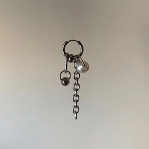Könnte beinhalten: Ein silberfarbener Creolen-Ohrring mit hängenden Anhängern. Das Design besteht aus einer kurzen Metallkette, einer kleinen polierten silberfarbenen Kugel und einer einzelnen weißen kugelförmigen Perle, die an einem runden Verschluss hängen.