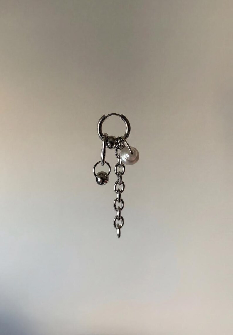 May include: A silver-toned hoop earring featuring a dangling charm assembly. The design includes a short metal chain, a small polished silver-colored sphere, and a single white spherical bead, all suspended from a circular hoop closure.